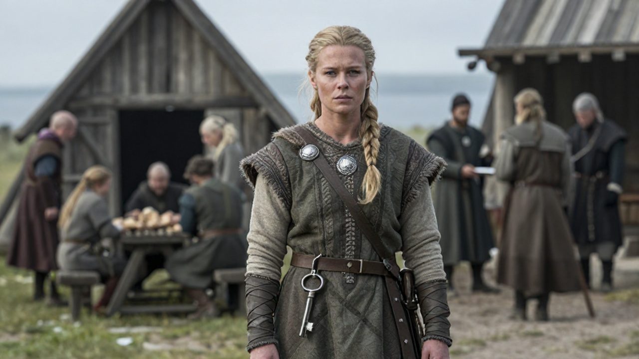Mujer vikinga con vestimenta tradicional y llaves de hierro al cinturón frente a un asentamiento nórdico.